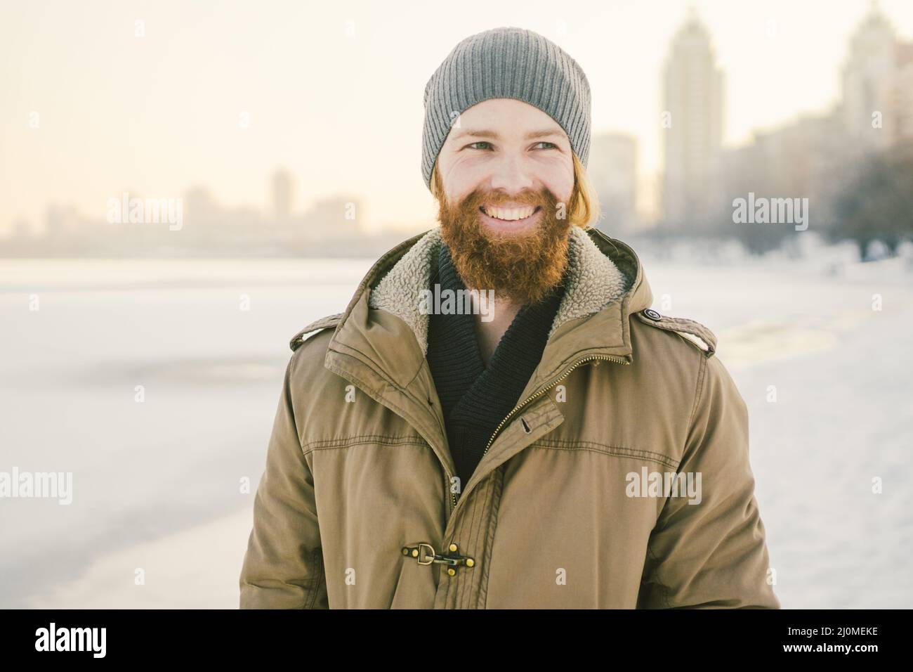 Close-up Caucasian young male red hair and beard in a hat and a park coat posing winter model against a background of a lake to Stock Photo