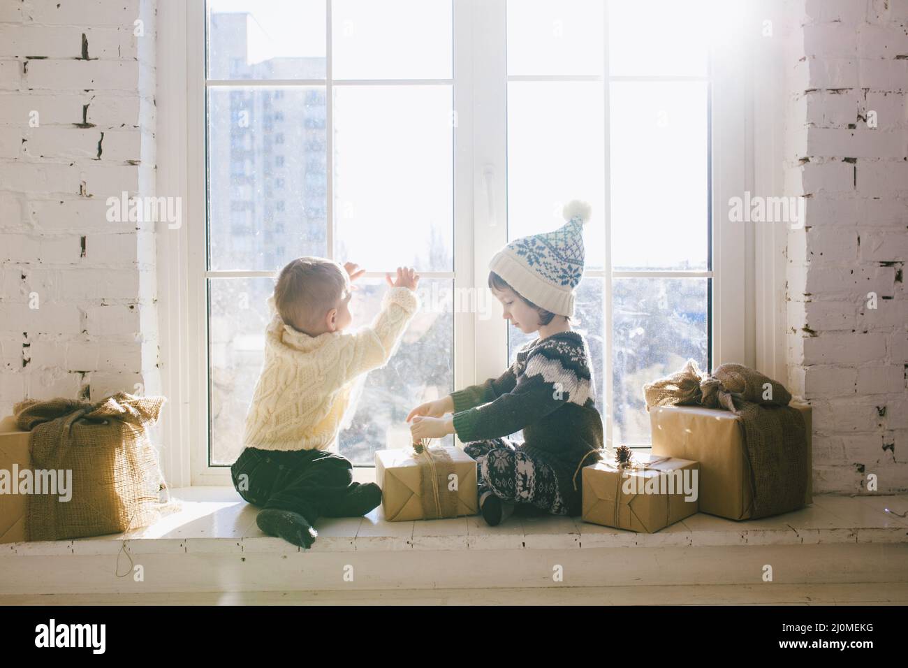 Children brother and sister of preschool age sit by window on a sunny Christmas day and play with gifts boxes wrapped in paper.T Stock Photo