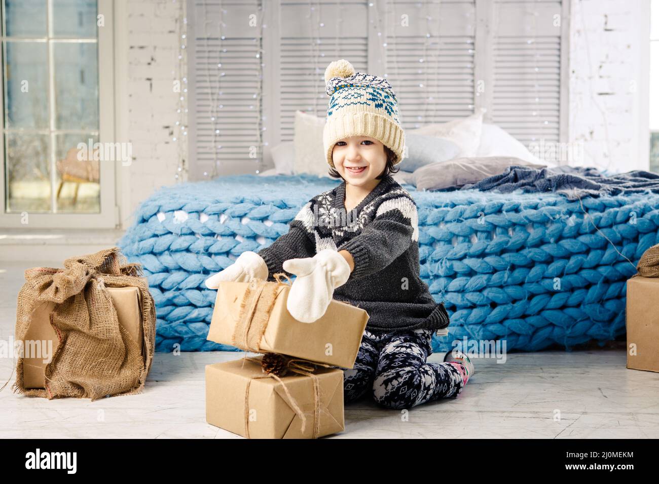 Little baby girl is folding up a mountain of gift boxes at home, near the bed. The interior is decorated with Christmas decor. D Stock Photo