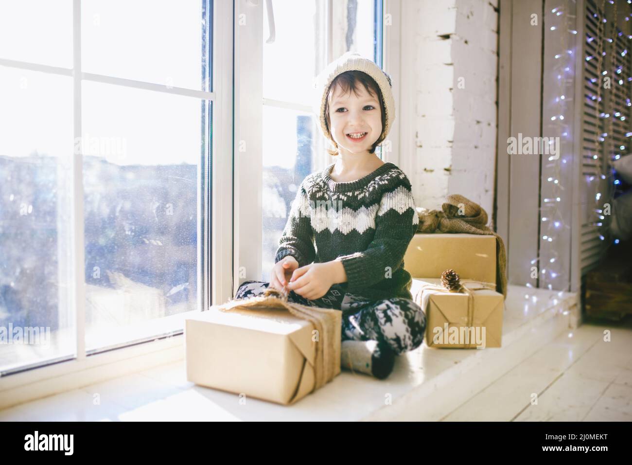 Child is sitting by the window on a sunny Christmas day and makes out with gifts in boxes wrapped in paper.dressed in knitted wa Stock Photo