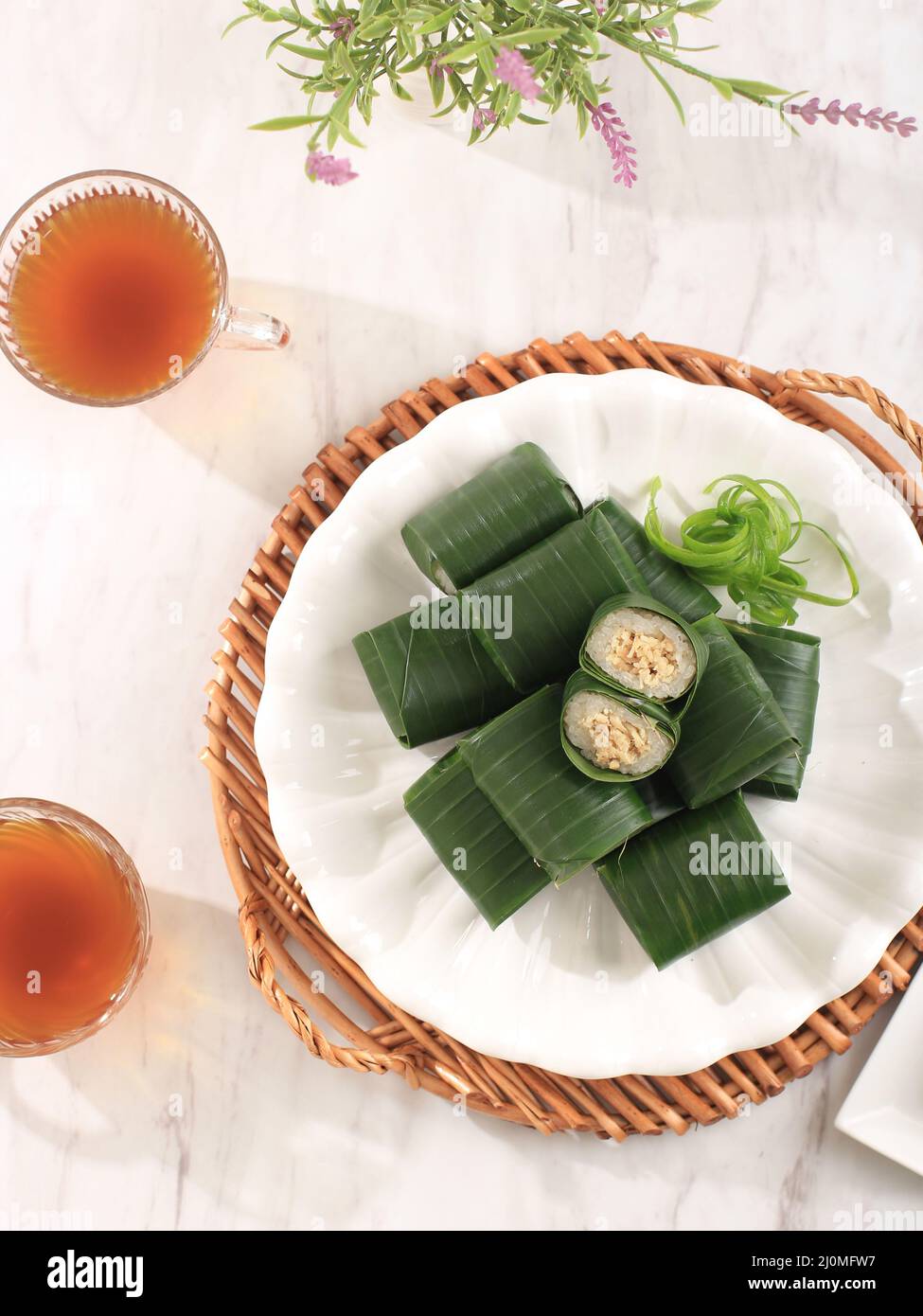 Top View Lemper Ayam, Steamed Glutinous Rice Filled with Shredded Chicken Wrapped in Banana Leaf on White Table. Served with Tea Stock Photo
