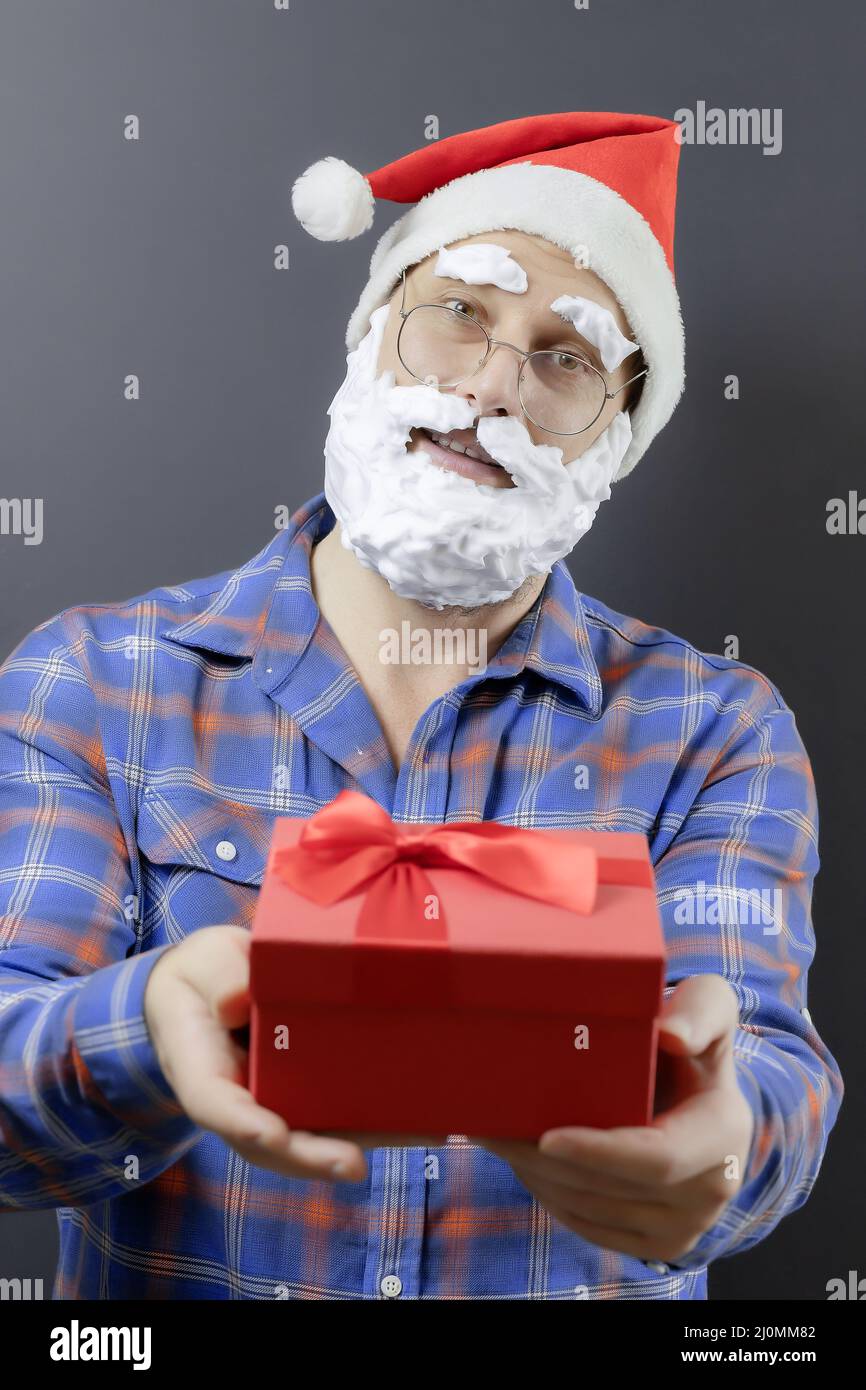 Santa in a plaid shirt with a white foam beard is holding a red gift box. Focus on the face. Merry Christmas to you Stock Photo