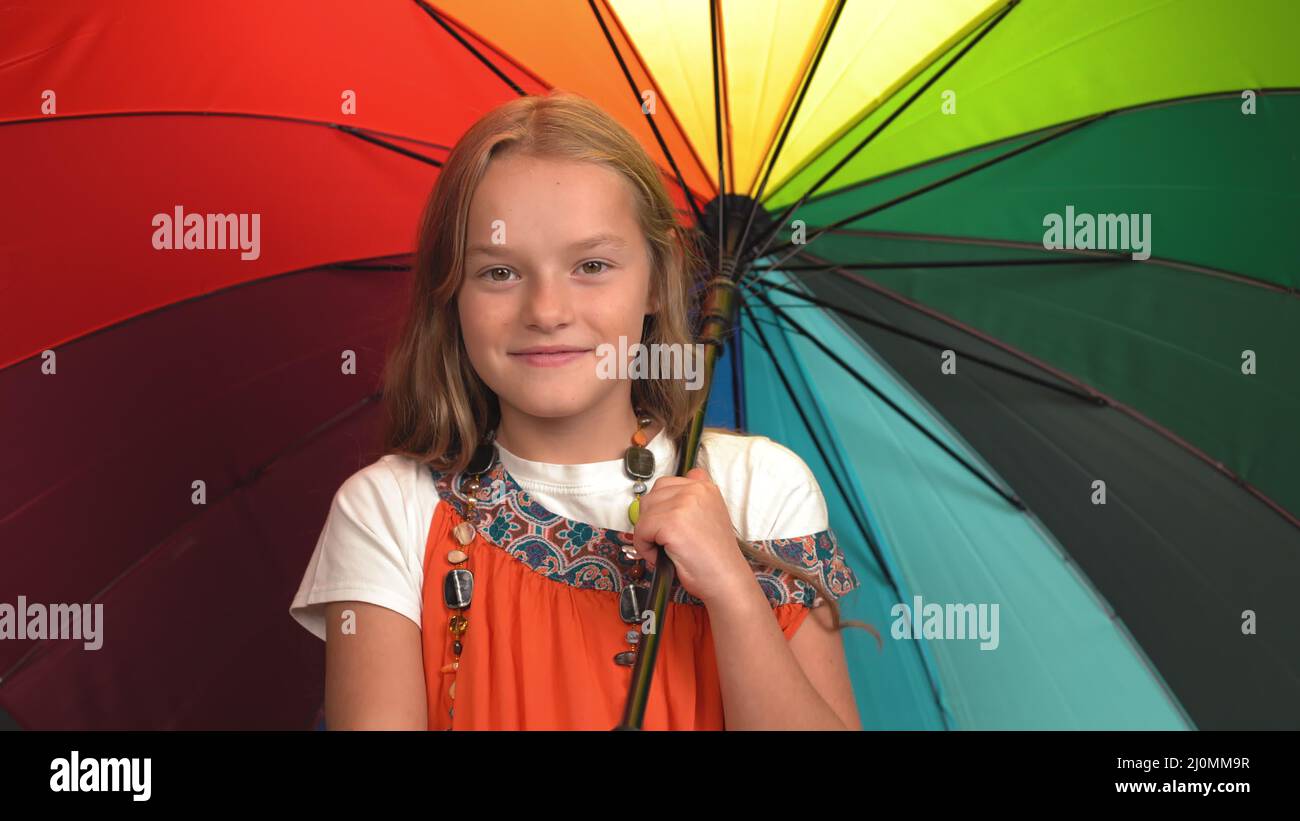 Smiling girl in mother's sundress with beads twists open bright multi-colored umbrella. Caucasian child looks at camera and smil Stock Photo