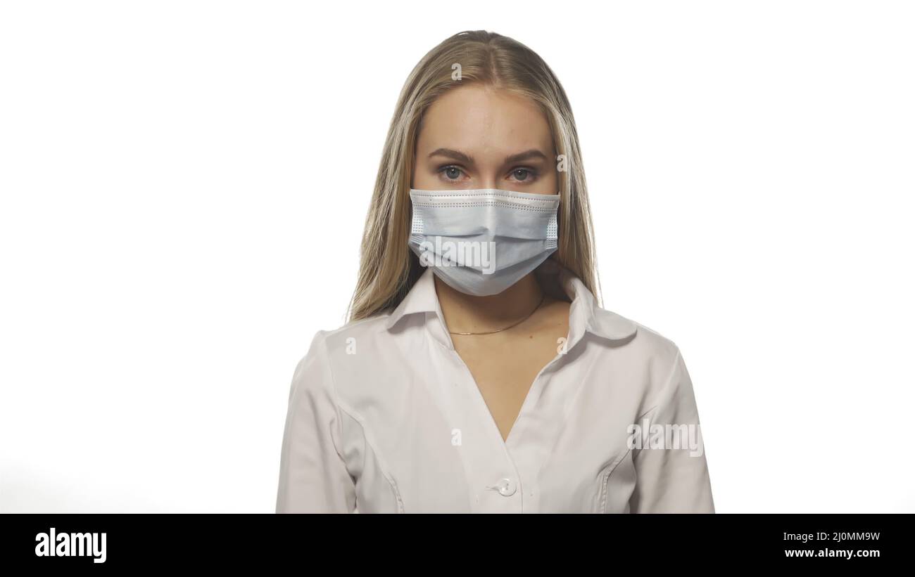 Blonde nurse in medical mask looks directly into camera on white background. Young woman with relief removes mask. Concept is en Stock Photo