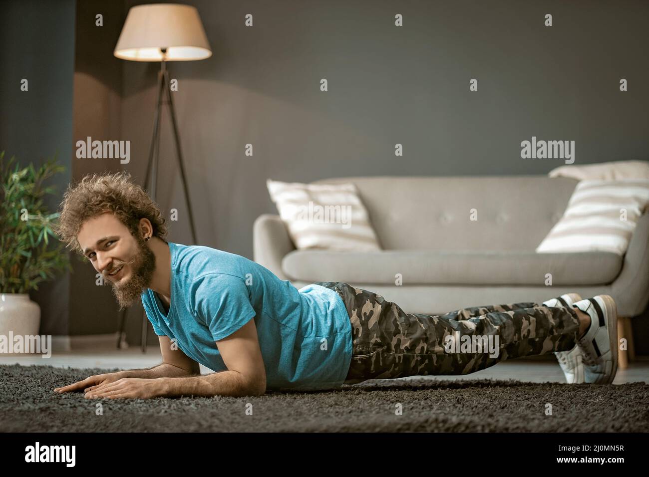 Cheerful Guy in a Blue T-shirt Does Plank Indoor. Young Man Does Up-downs to Get Fit. Medium close-up Stock Photo