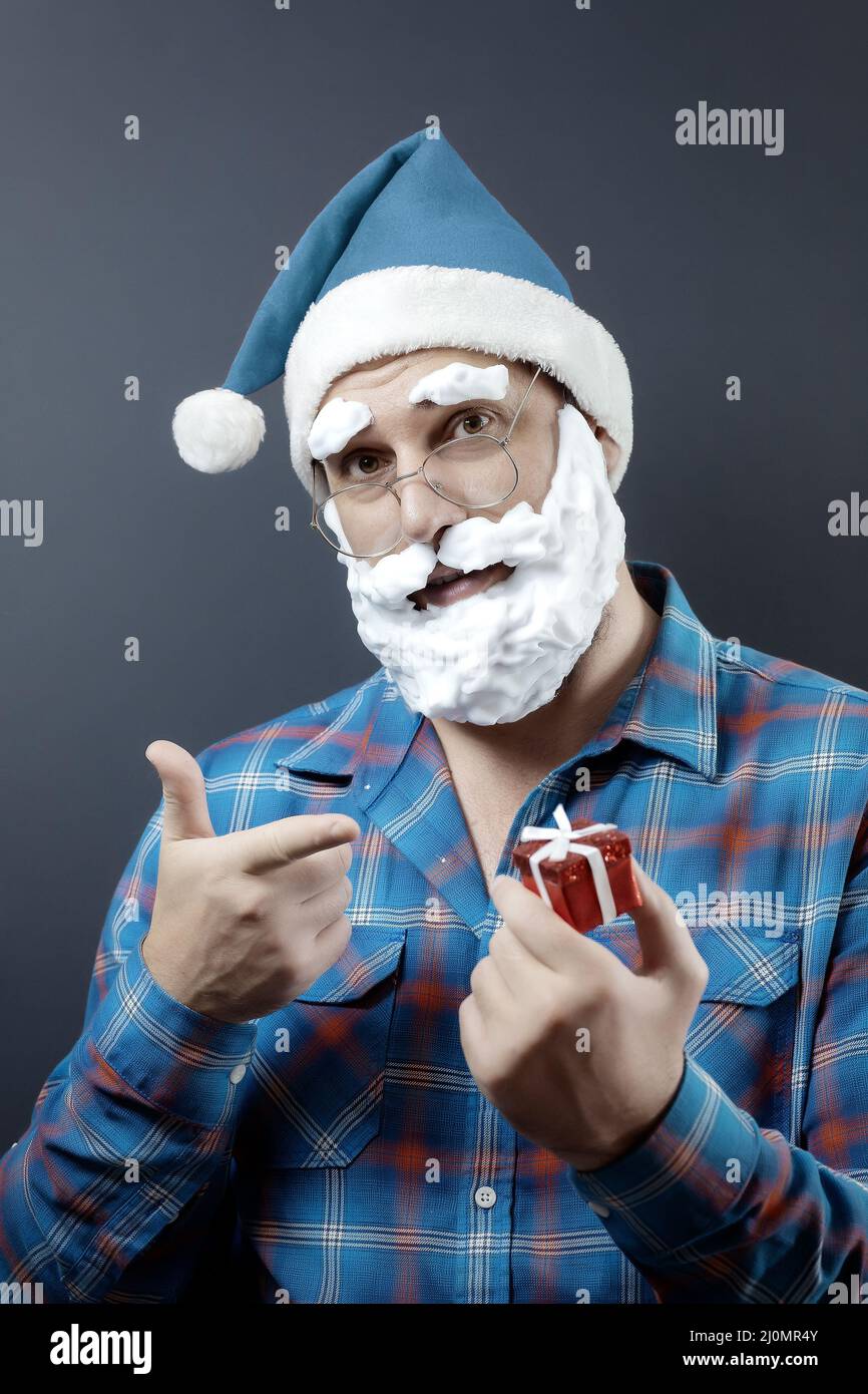 Santa with a foam beard holds a tiny red gift box and points his index finger at it. Hand with a gift is defocused Stock Photo