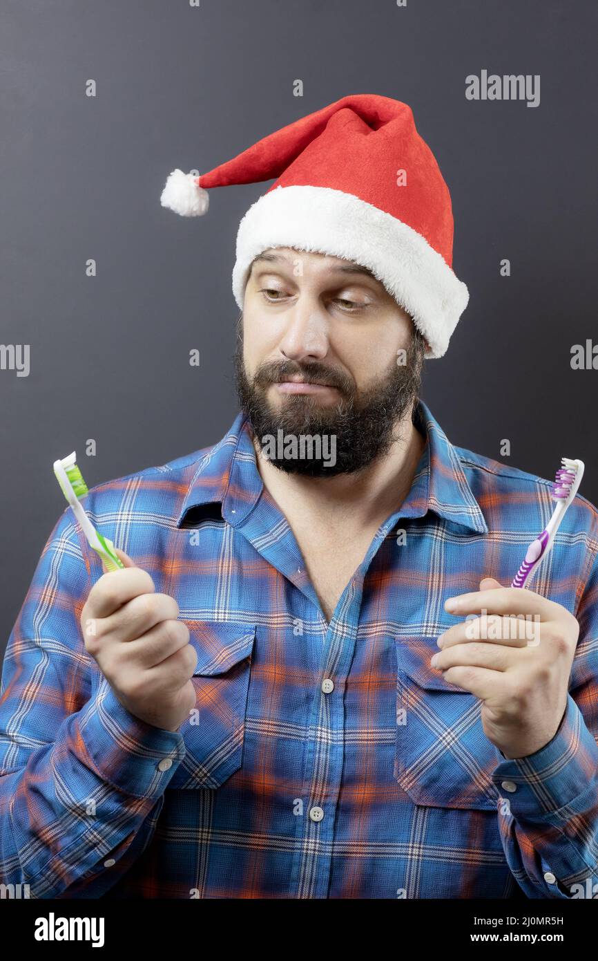 A pensive bearded man in a Santa hat is choosing from two toothbrushes. Preparing for Christmas. Fake santa Stock Photo