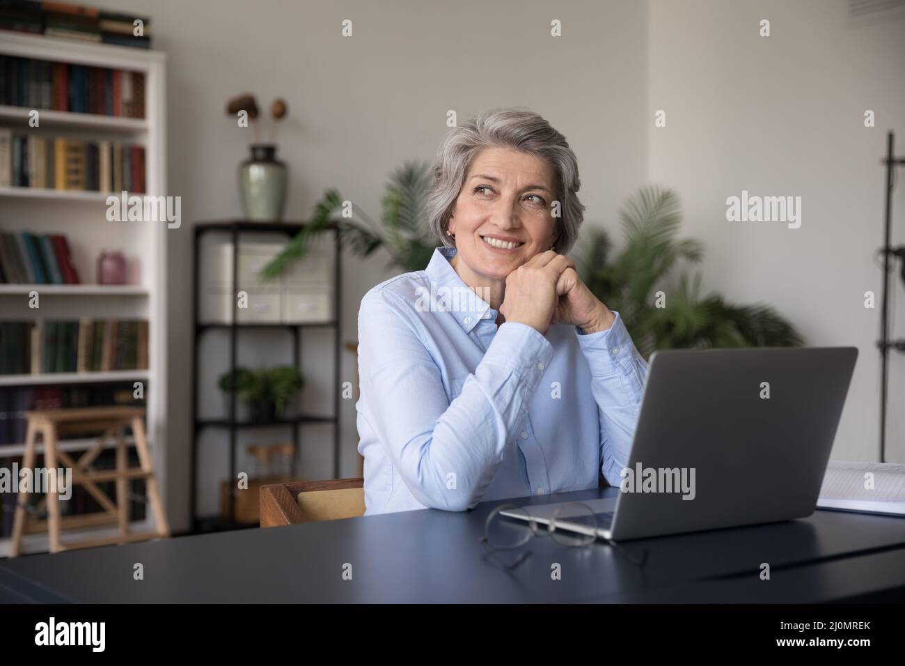 Happy older woman sit at workplace desk looking into distance Stock Photo