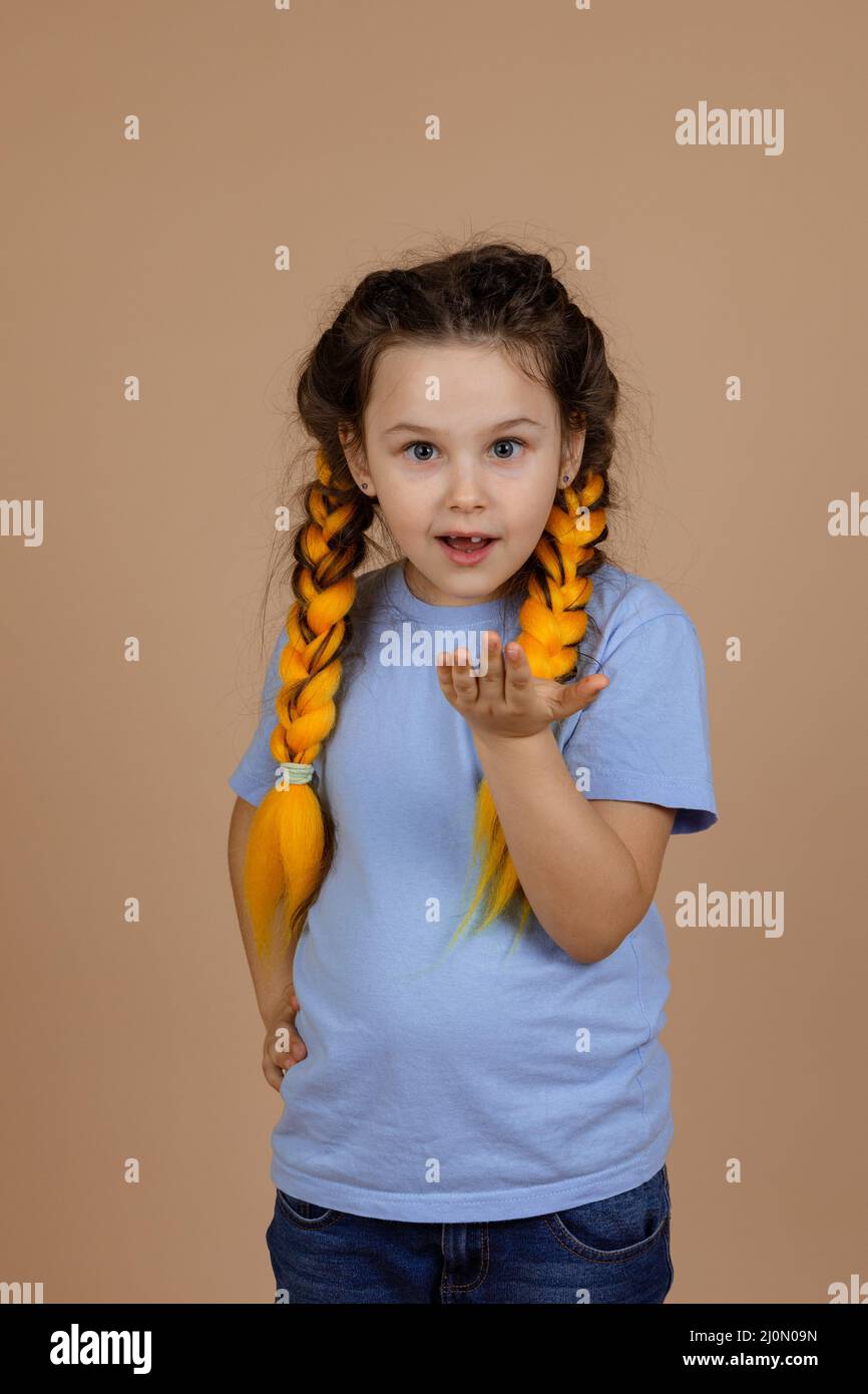 Surprised young girl asking something showing hand looking at camera with yellow kanekalon braids on head on beige background wearing blue t-shirt and Stock Photo