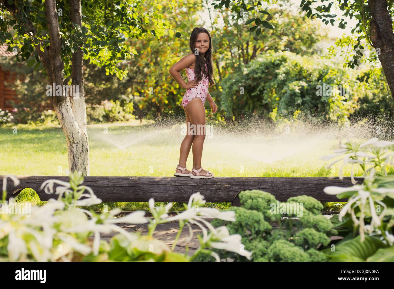 Small playing girl on log smiling looking at camera balancing indulging with water sprinklers, house and green trees in background in daytime Stock Photo
