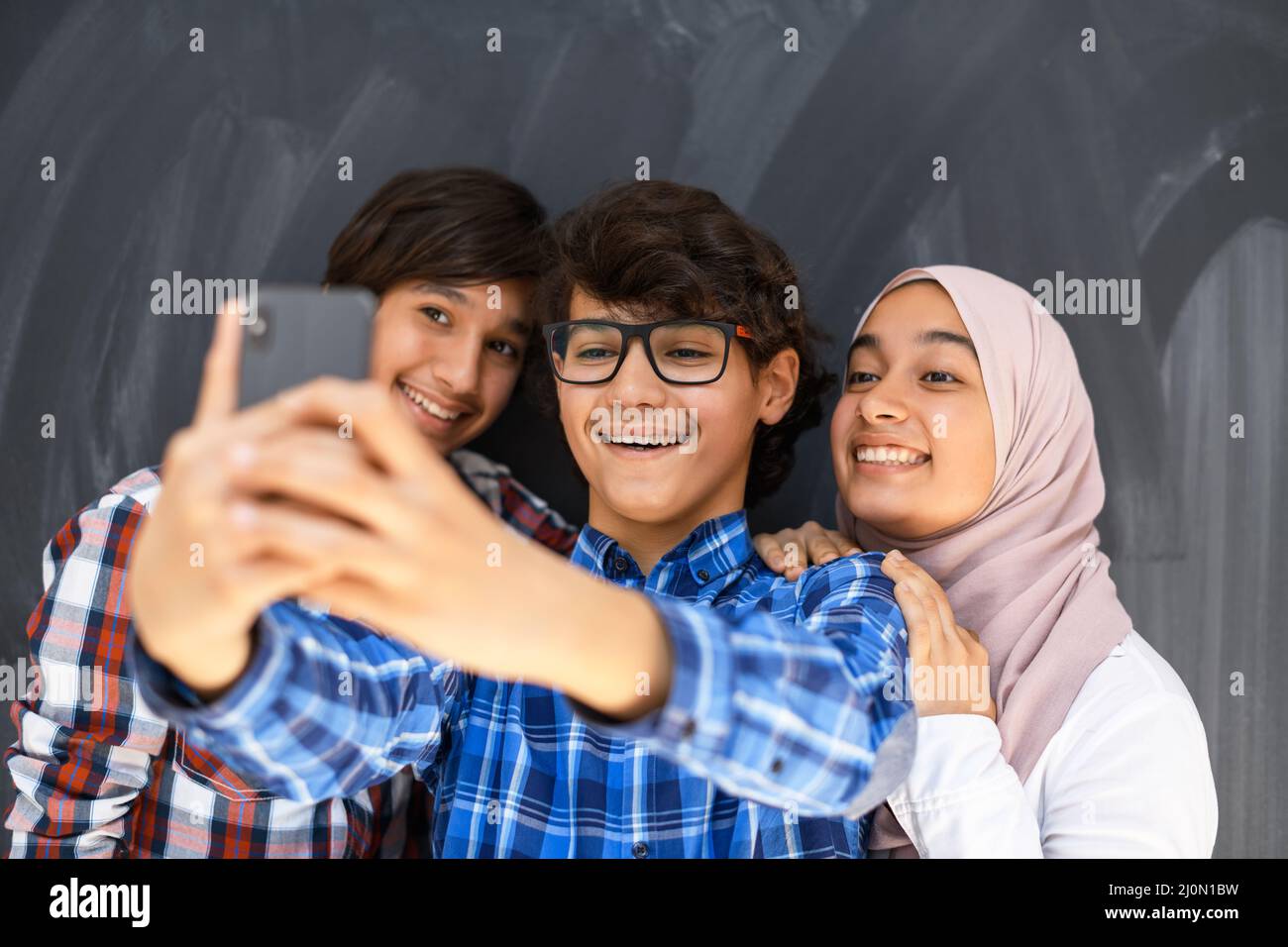 Group of arab teens taking selfie photo on smart phone with black chalkboard in background. Selective focus Stock Photo