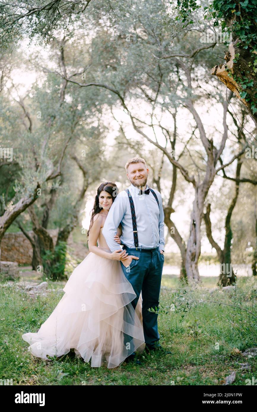 The bride and groom are standing among the trees in the olive grove ...