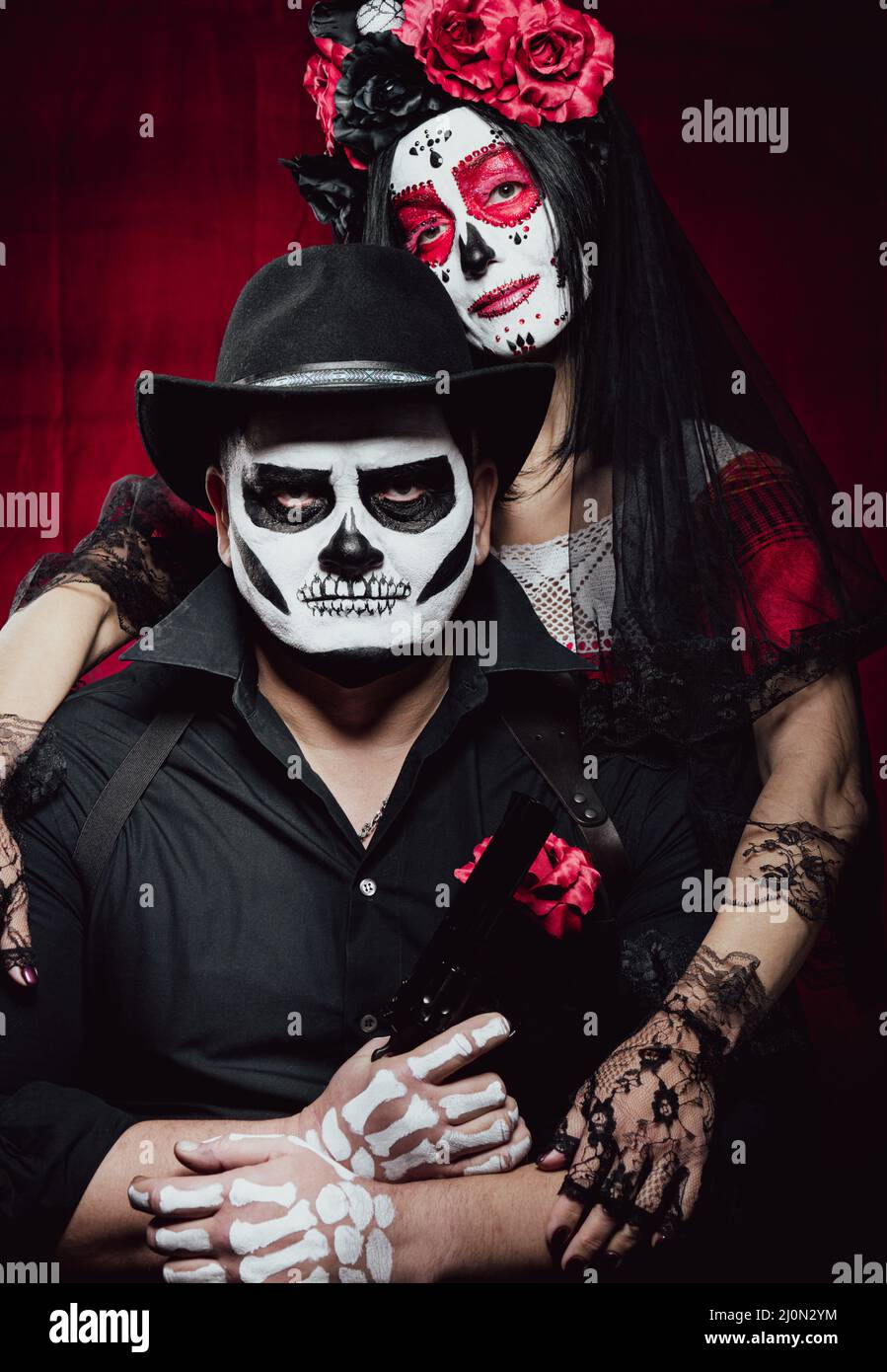 Beautiful woman with a sugar skull makeup with a wreath of flowers on her head and a skeleton man in a black hat holding a gun Stock Photo
