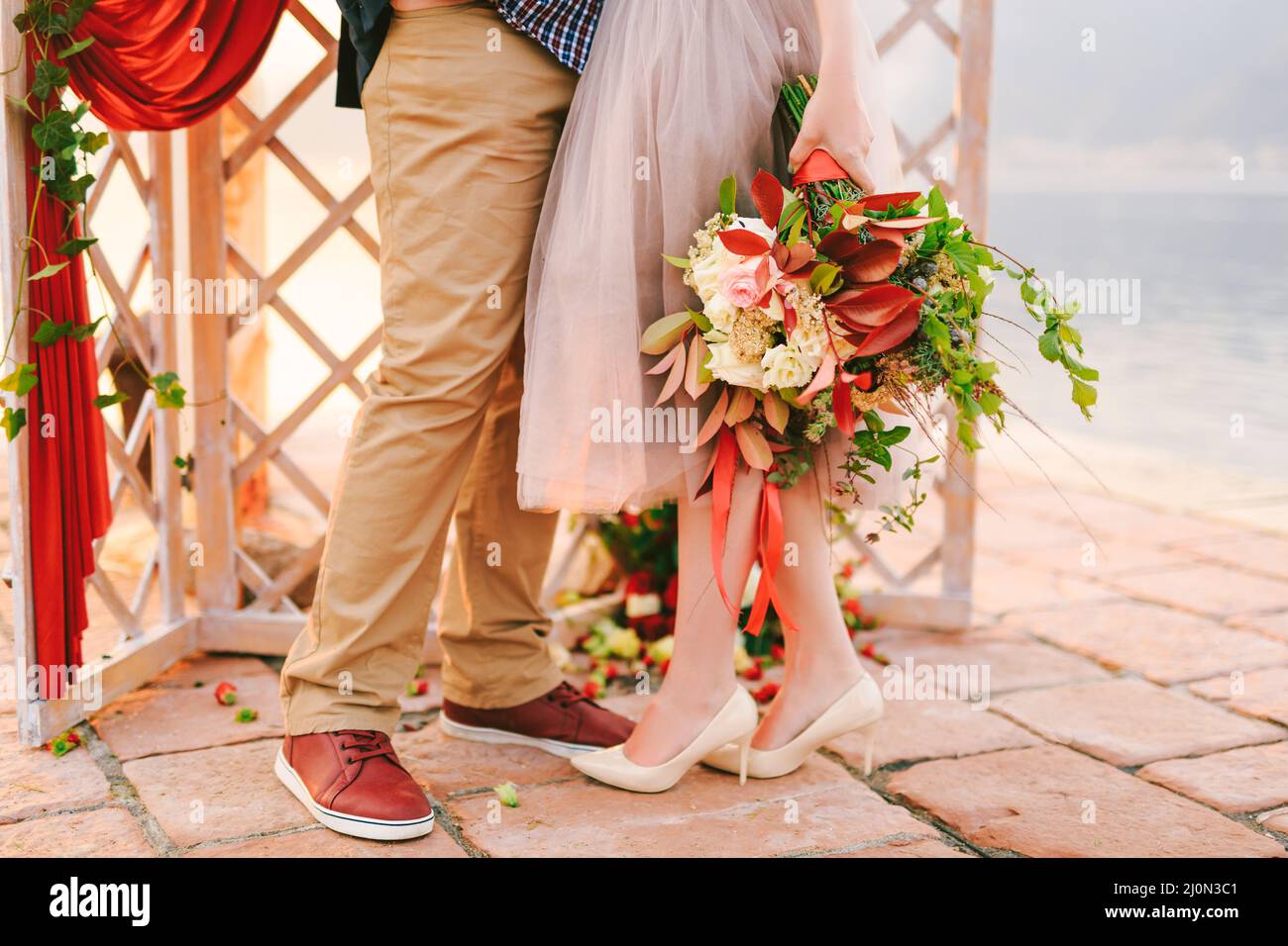 Groom and bride with a bouquet of flowers stand on a stone tile against the background of a carved screen decorated with a red c Stock Photo