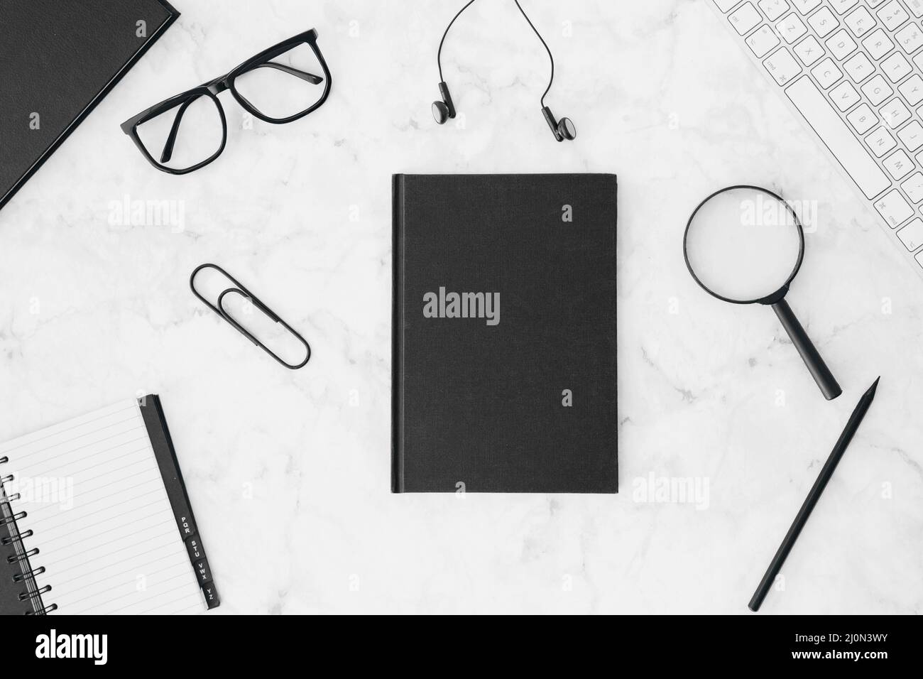 Closed black diary with eyeglasses earphone magnifying glasses pencil keyboard diary white desk Stock Photo