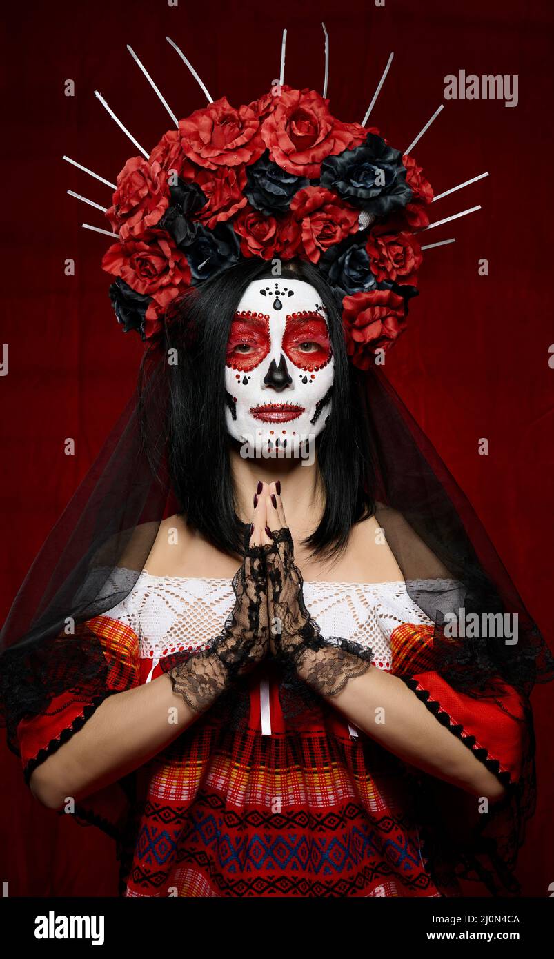 Beautiful woman with a sugar skull makeup with a wreath of flowers on her head and a skull hands raised up in prayer pose in bla Stock Photo