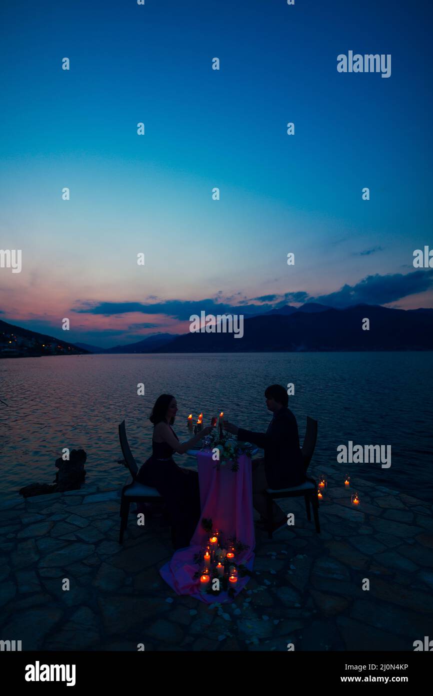 Man and woman sit at a table and clink glasses on the pier against the background of the sea and mountains at night by burning c Stock Photo