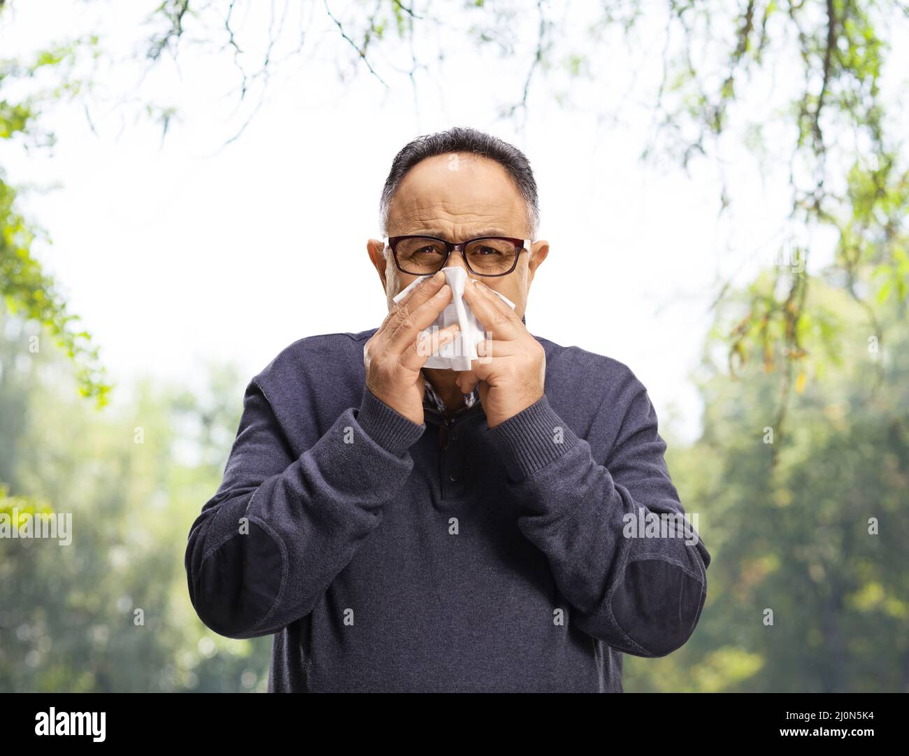 Mature man blowing nose with a paper tissue in a park with trees behind Stock Photo