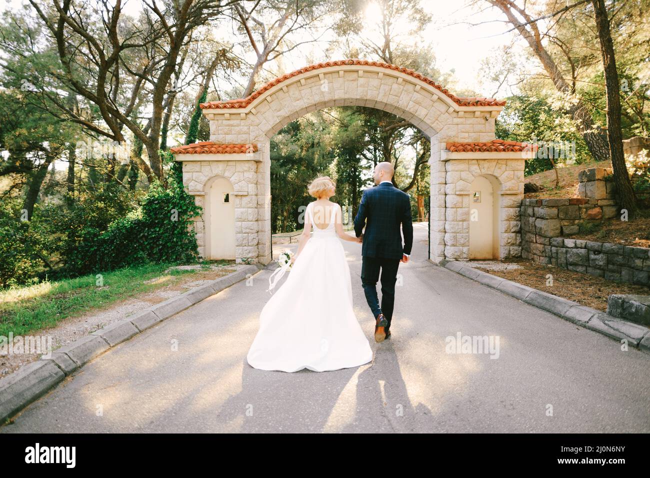 Newlyweds walk along the path holding hands to the entrance stone gate with orange tiles in a green park. Back view Stock Photo