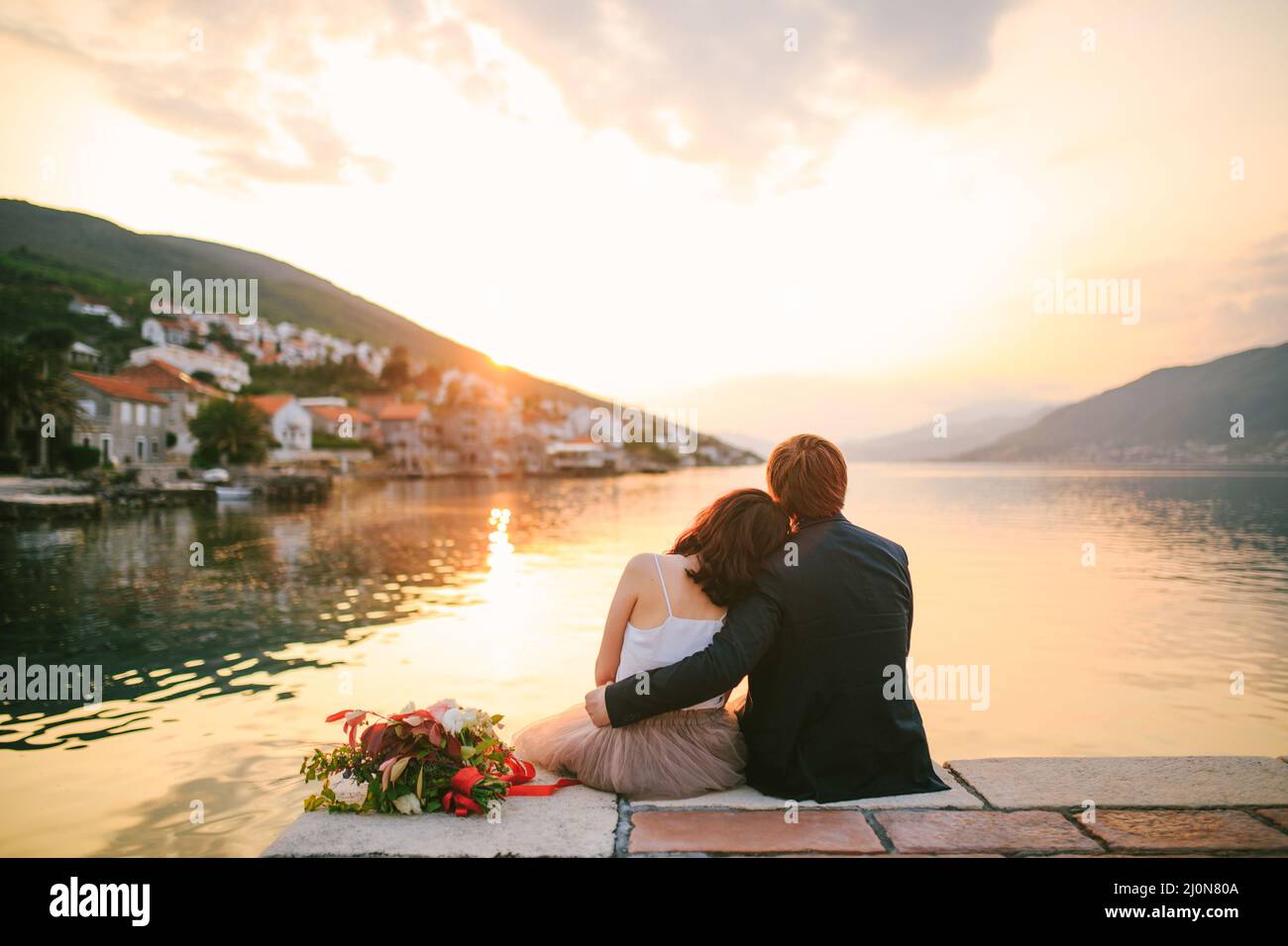 Groom hugs the bride, who has laid her head on his shoulder, sitting on the coast against the background of mountains, sea, old Stock Photo