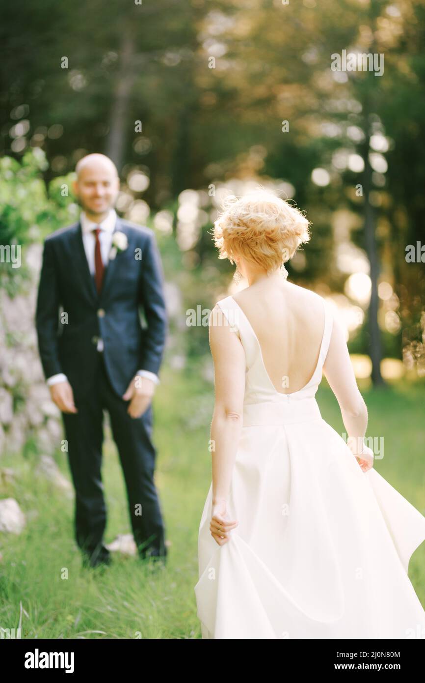 Bride in a white dress is walking towards smiling groom against the background of green trees Stock Photo