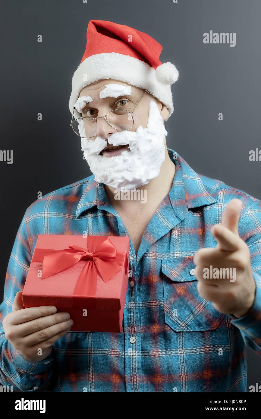 Santa in a plaid shirt with a white foam beard holds a red gift box and points his finger. The hand is out of focus. Gift for yo Stock Photo