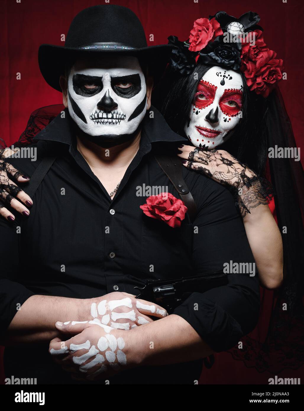 Beautiful woman with a sugar skull makeup with a wreath of flowers on her head and a skeleton man in a black hat. Couple on dark Stock Photo
