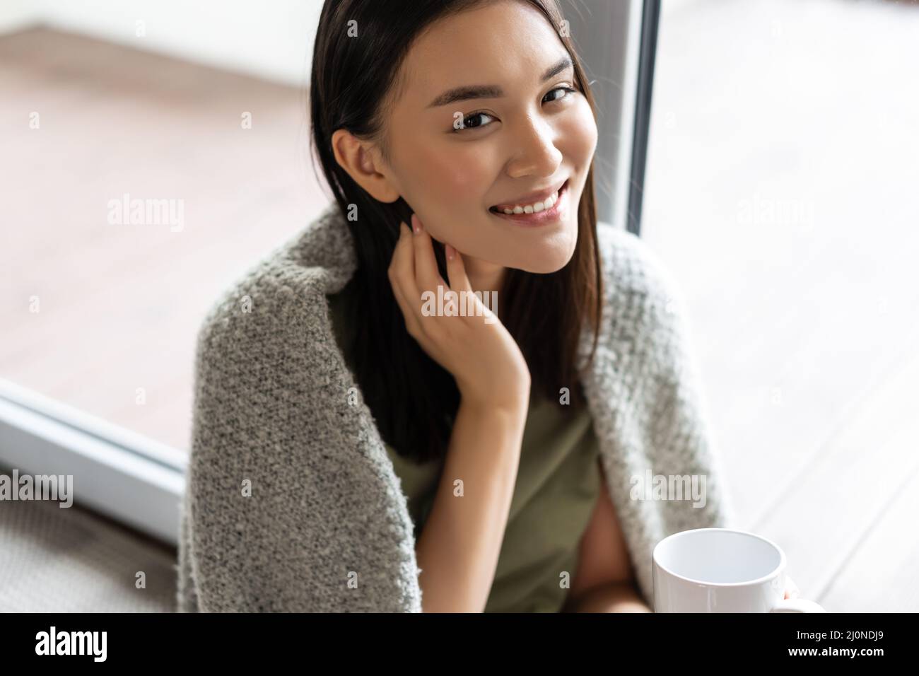 Portrait of beautiful asian woman wrapped in blanket, drinking coffee and smiling, relaxing at home on weekend, sitting near win Stock Photo