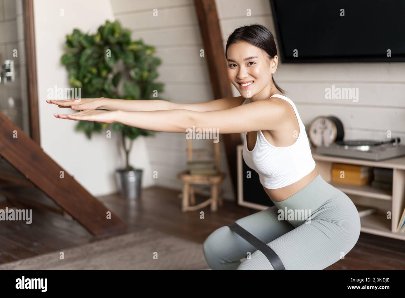 Cute asian fitness girl at home doing workout, squats with stretching elastic rope on her legs, exercising for fit and healthy b Stock Photo