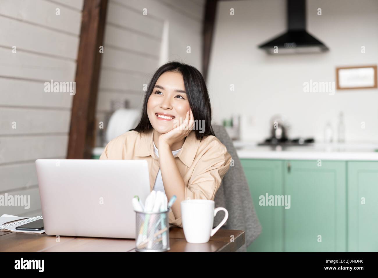 Smiling asian woman working on laptop from home, dreaming of something with happy face, thinking while sitting near computer Stock Photo