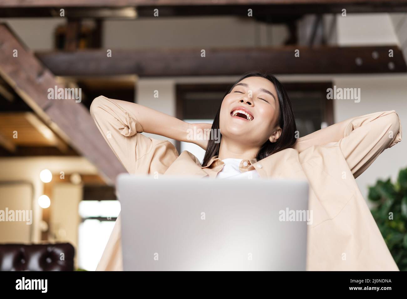 Smiling asian girl relaxing with laptop at home, lay back with hands behind head and pleased face expression Stock Photo