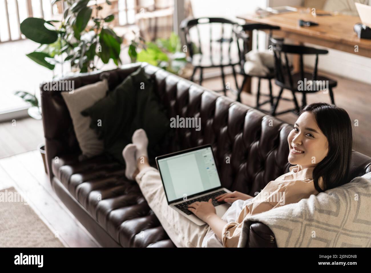 Smiling asian woman working freelance from home, resting on couch with laptop computer, looking happy at camera Stock Photo