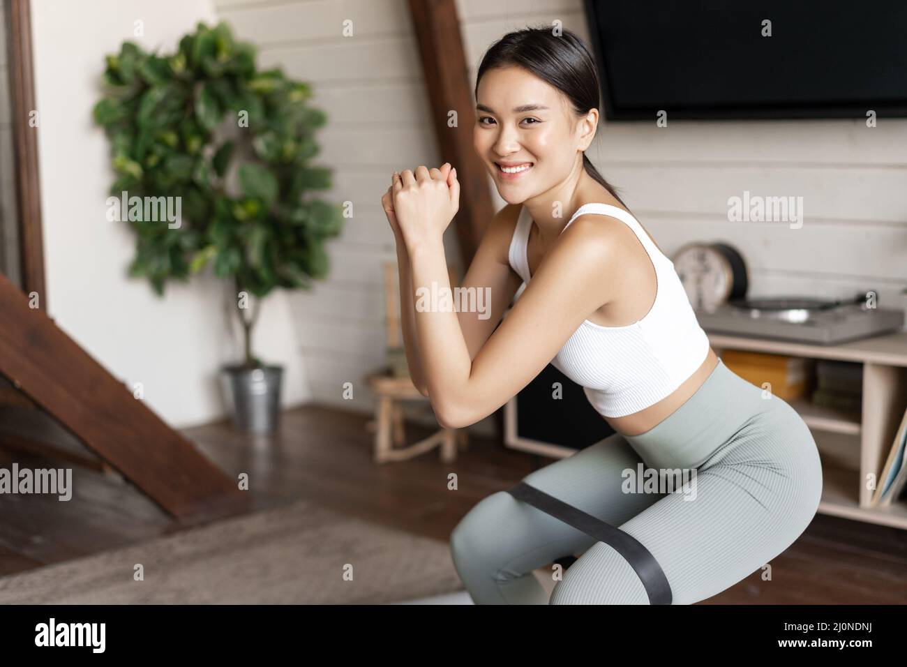 Cute asian fitness girl at home doing workout, squats with stretching elastic rope on her legs, exercising for fit and healthy b Stock Photo