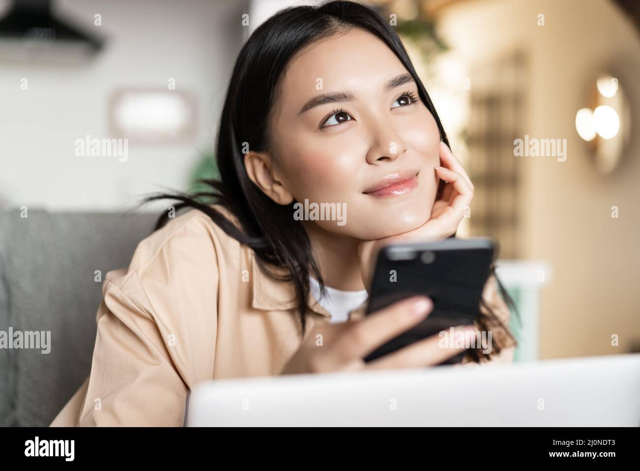 Smiling asian girl holding phone and thinking, looking up dreamy, sitting with laptop at home Stock Photo