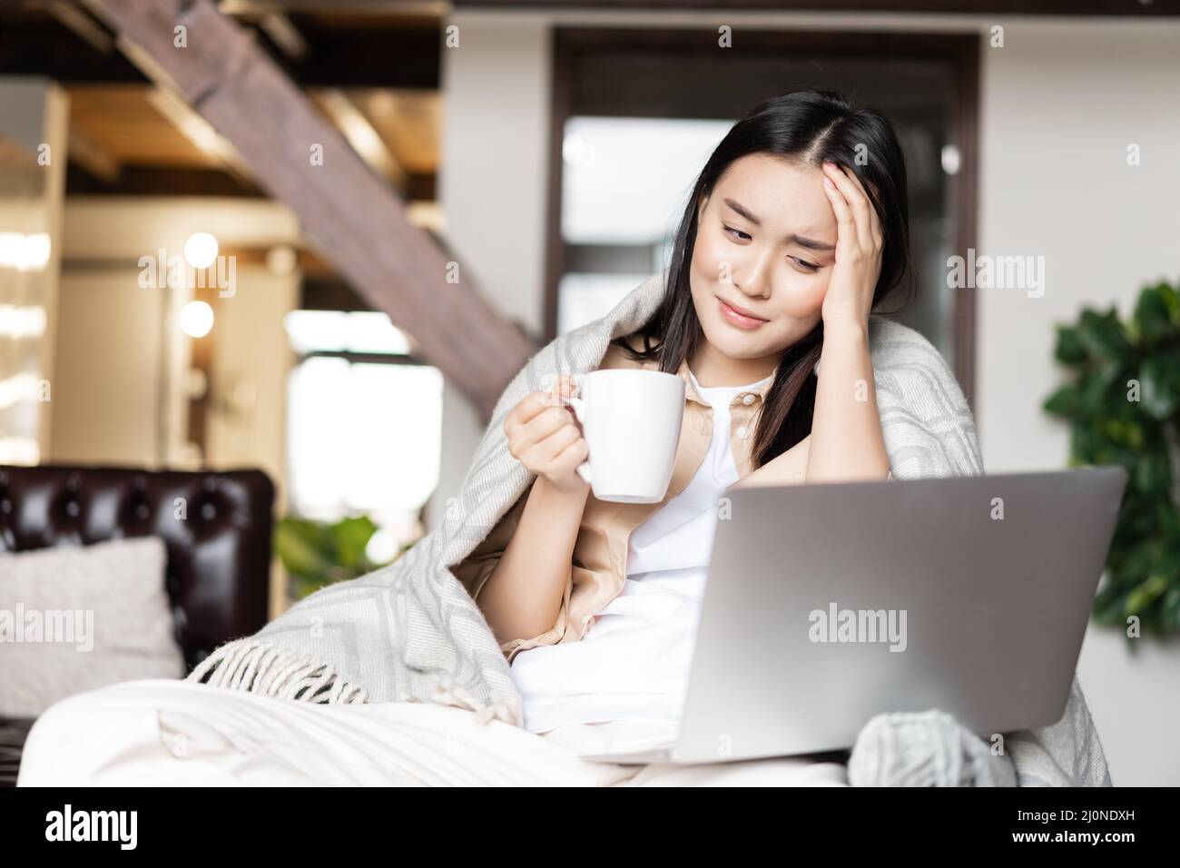 Young sick asian girl staying at home on quarantine, catching flu or covid, watching videos on laptop and drinking tea Stock Photo