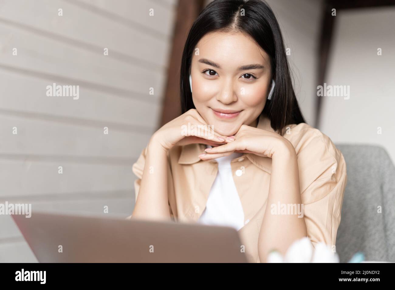 Portrait of asian woman in earphones working on laptop and smiling, looking happy at camera, studying or learning online course Stock Photo