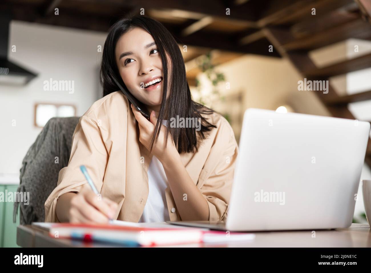 Young professional asian woman working from home, answer phone call, talking to client and writing down information, taking note Stock Photo