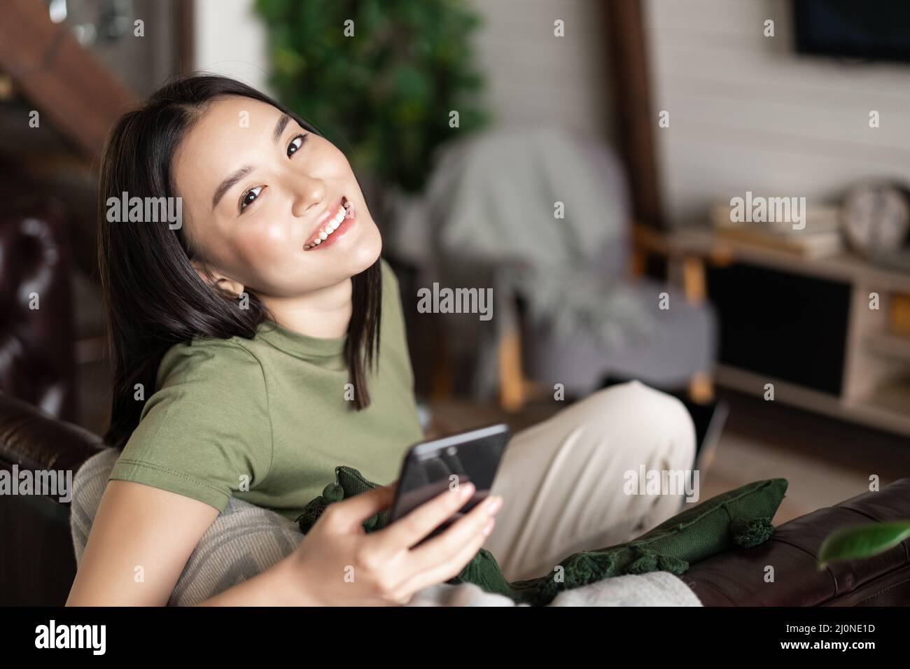 Dreamy smiling asian woman using mobile phone, sitting and relaxing at home Stock Photo