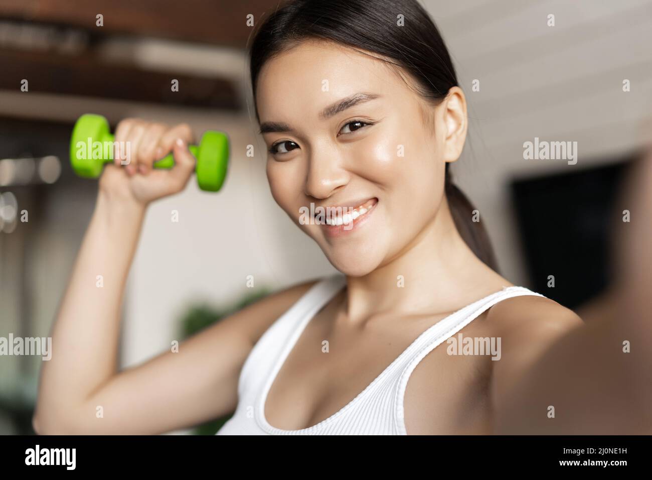 Portrait of smiling asian fitness instructor taking selfie with dumbbell. Athletic woman making photo on mobile phone with sport Stock Photo