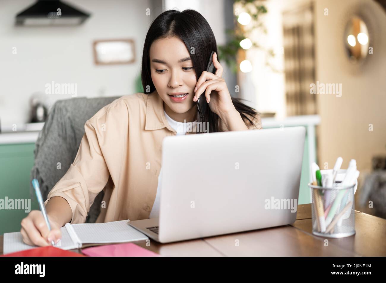 Asian woman working from home, answer phone call and writing down in notebook. Girl entrepreneur busy on cellphone, sitting near Stock Photo