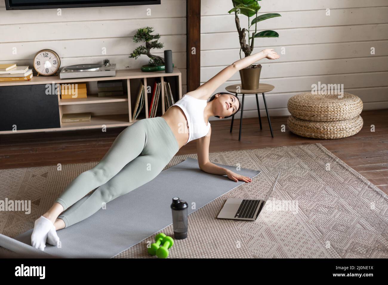 Young asian woman doing workout at home on floor mat, following sport instructor on laptop video, stretching arm and doing plank Stock Photo