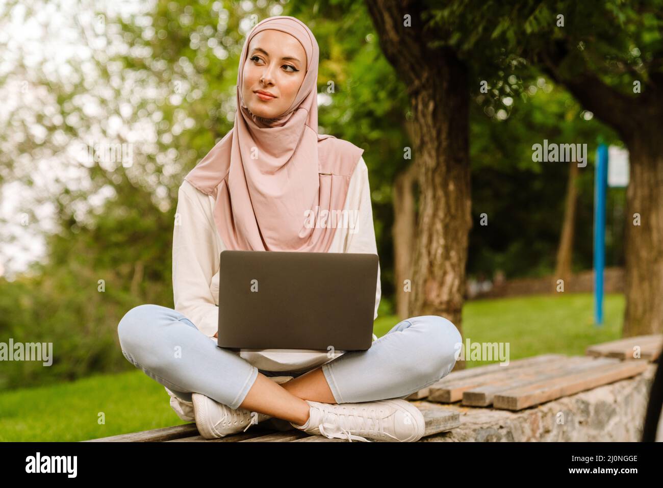 Happy young muslim woman in hijab sitting on a bench with laptop ...