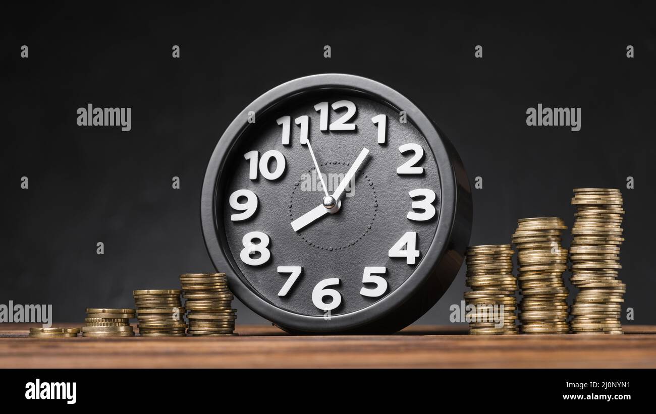 Round clock between the increasing coins on wooden desk against black ...
