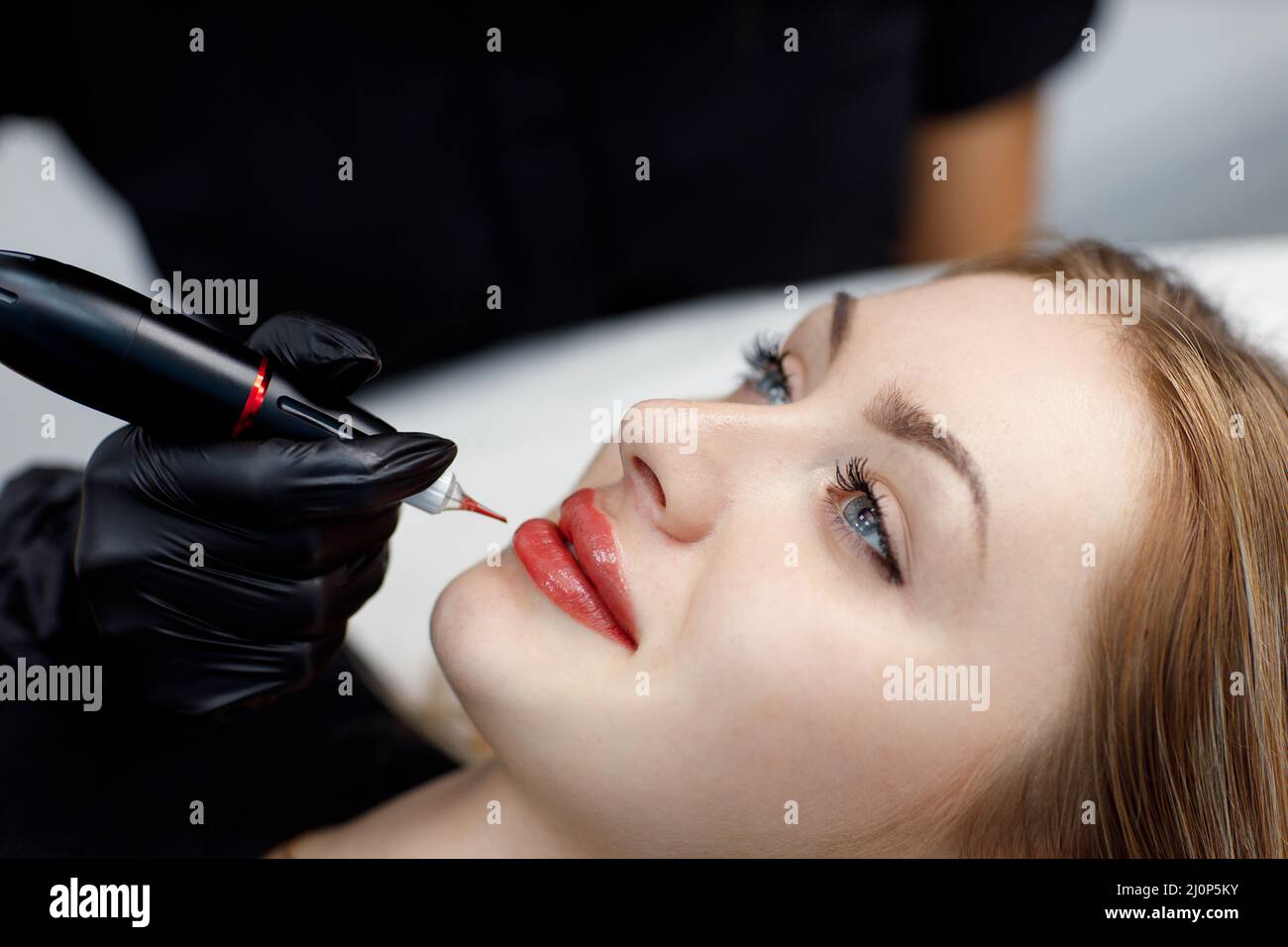 Cosmetologist applying pink pigment permanent tattoo on female lips with tatooing needle machine. Stock Photo