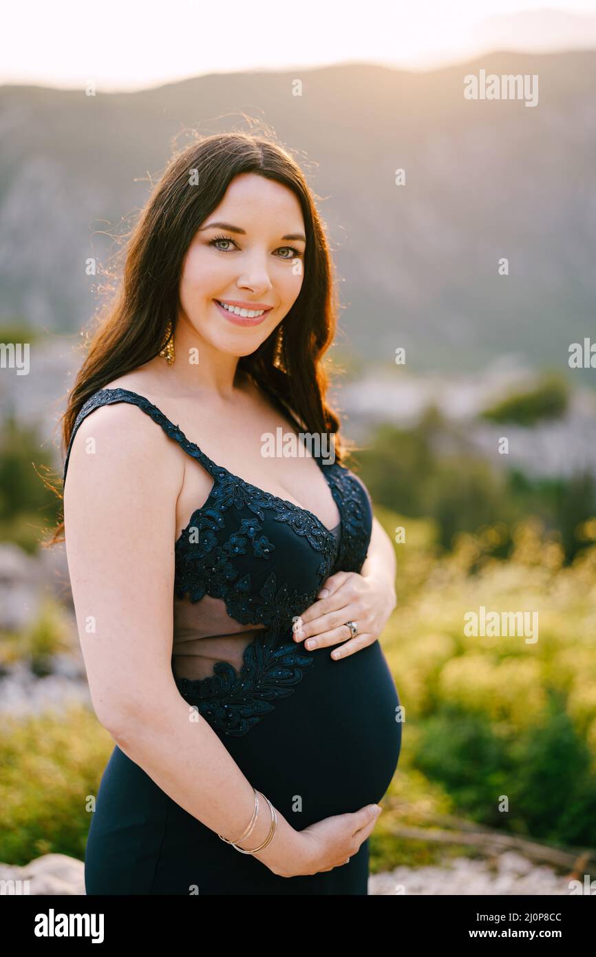 Pregnant woman hugging her tummy is standing on the top of a rocky mountain Stock Photo