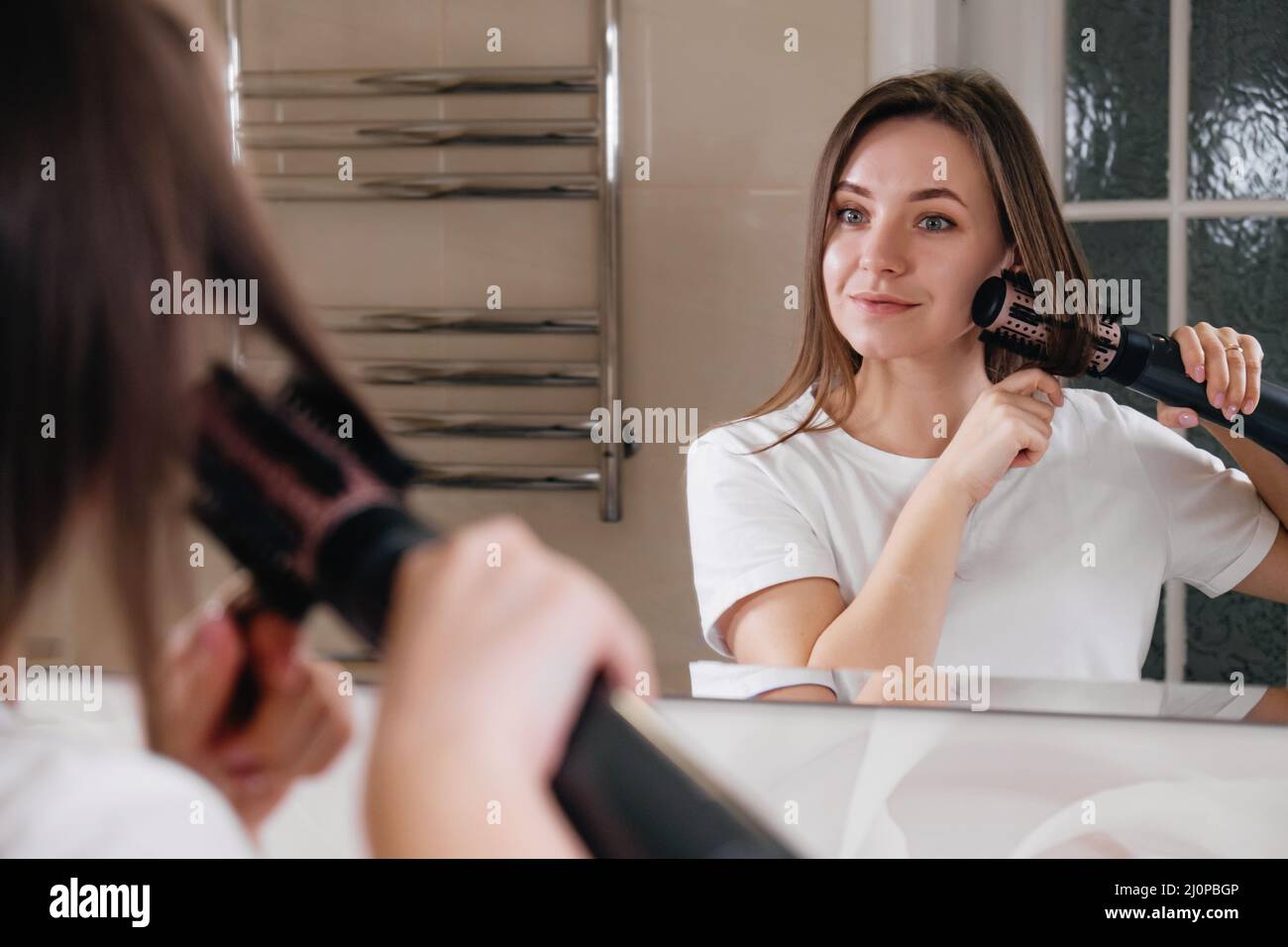 Woman drying her hair, styling in the bathroom, looking at the mirror, combing hair at home Stock Photo