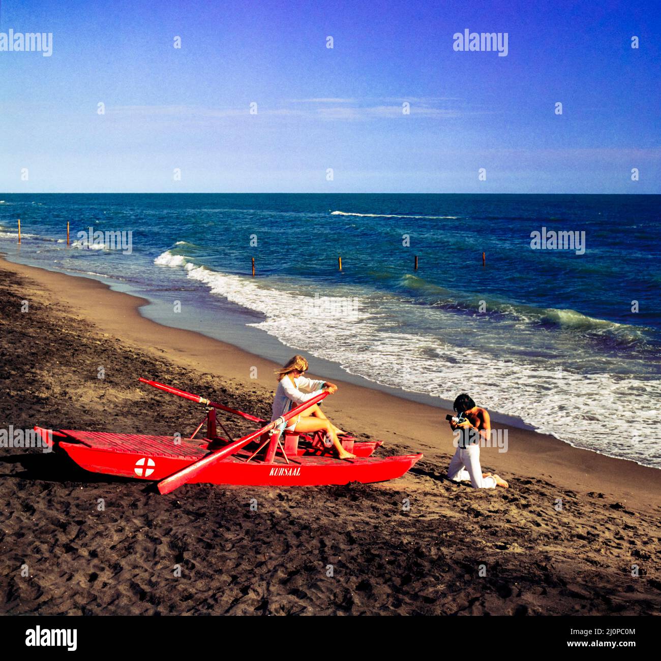 Vintage Italy 1970s, photographer shooting pictures, female fashion model, red rowing rescue boat, Kursaal beach, Lido di Ostia, Lazio, Europe, Stock Photo
