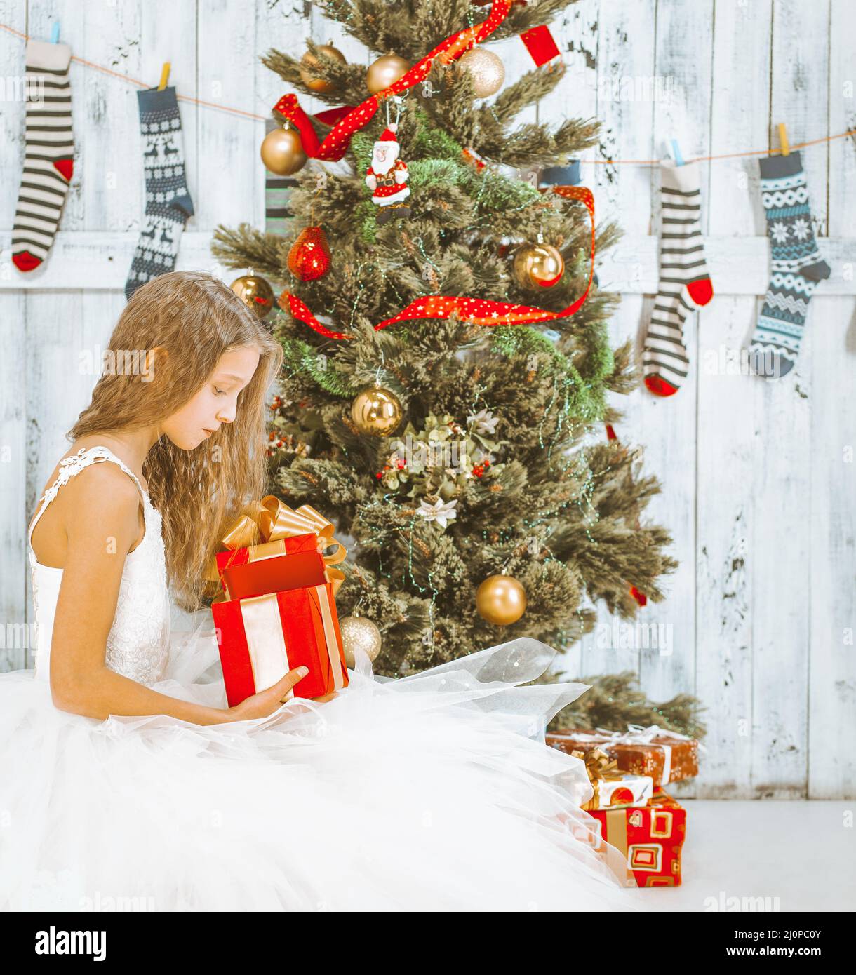 Teenage Girl with Long Hair in a White Tutu Dress Sits under a Christmas Tree and Unpacks her Present. Christmas Morning Stock Photo