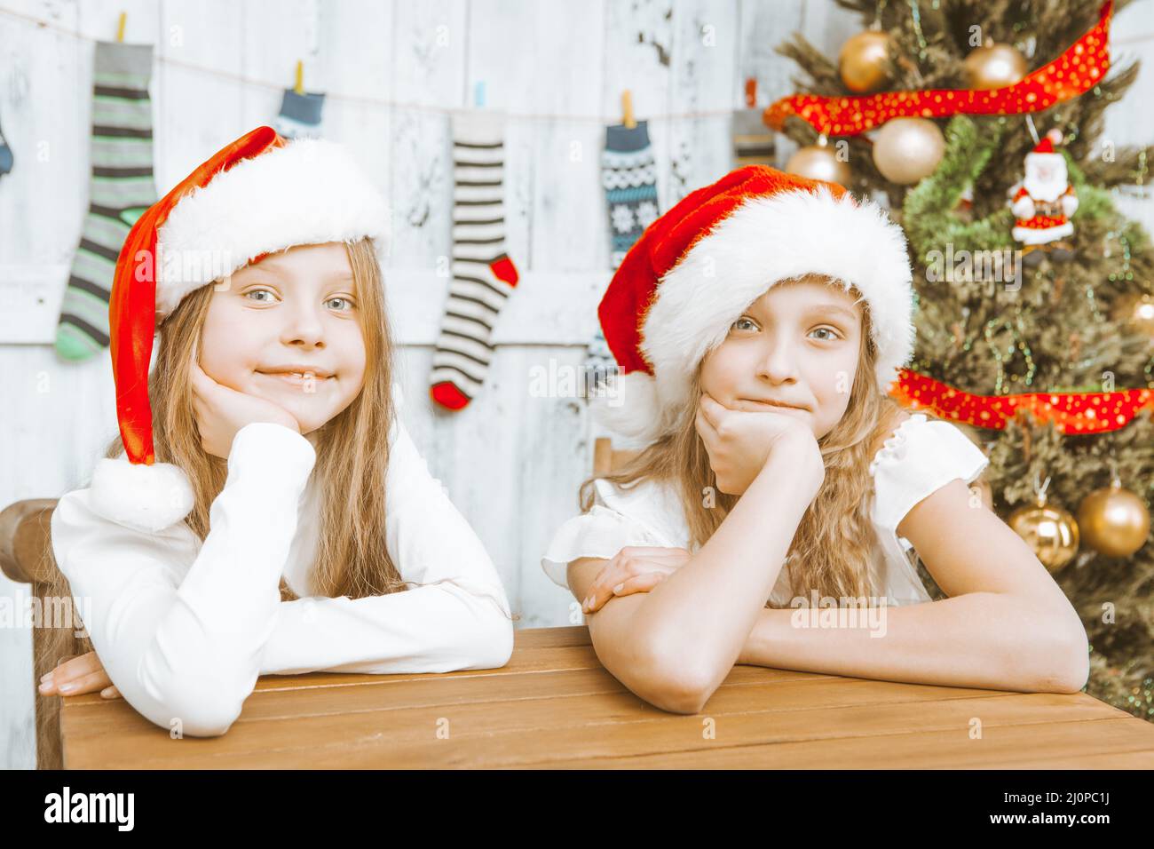 Children Sit at the Table Next to the Christmas Tree and Christmas Miracle and Gifts. Sisters in Santa Hats in the Christmas Mor Stock Photo
