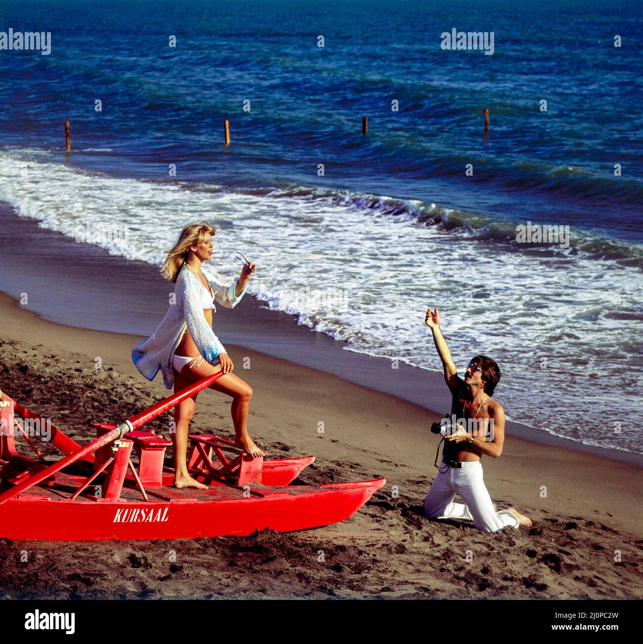 Vintage Italy 1970s, photographer shooting pictures, female fashion model, red rowing rescue boat, Kursaal beach, Lido di Ostia, Lazio, Europe, Stock Photo